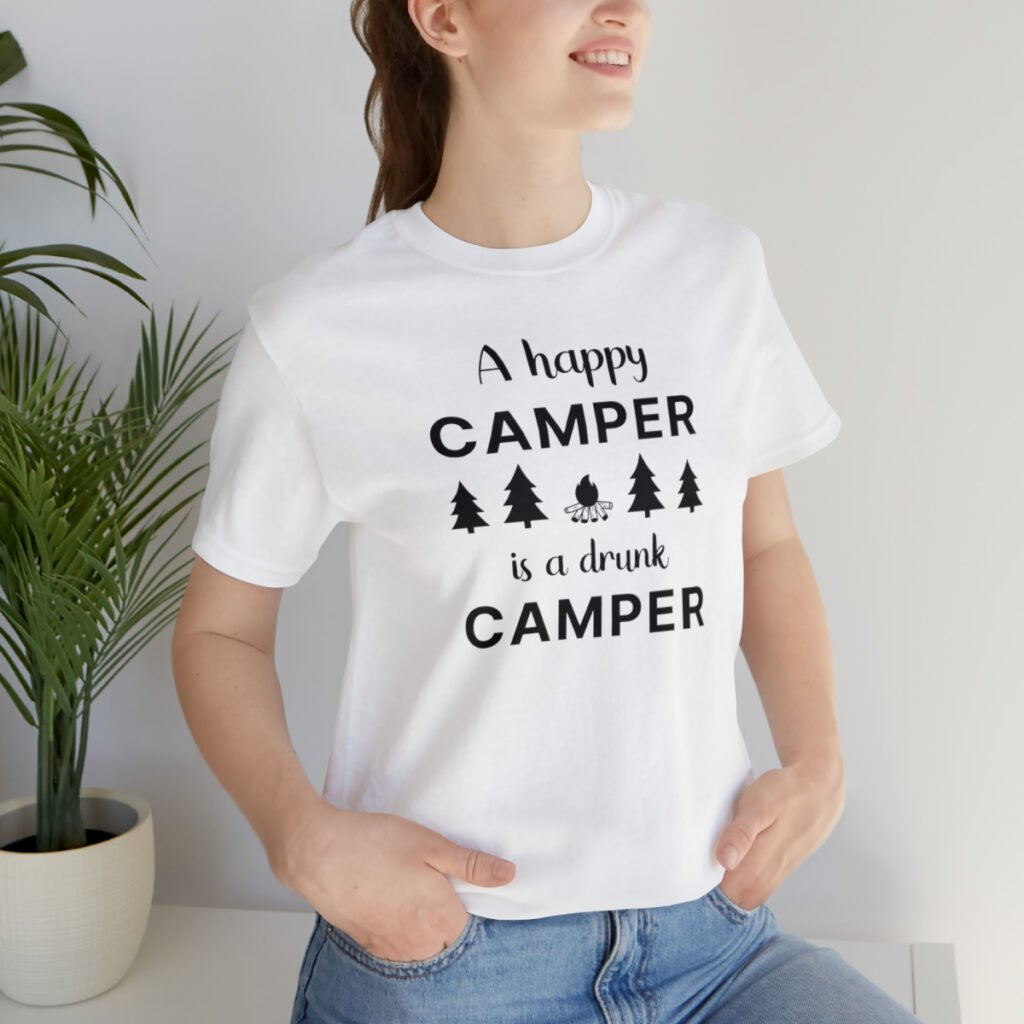 A young woman wearing a white Funny T-Shirt with an illustration of fur trees and a campfire saying "A happy camper is a drunk camper".