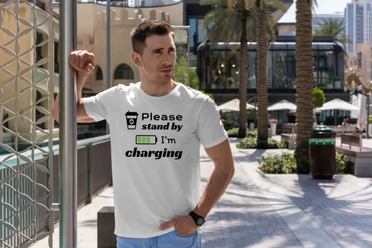 A handsome man outside wearing a white Funny T-Shirt for Coffee Lovers featuring a picture of a coffee cup and a battery saying "Please, stand by. I'm charging".