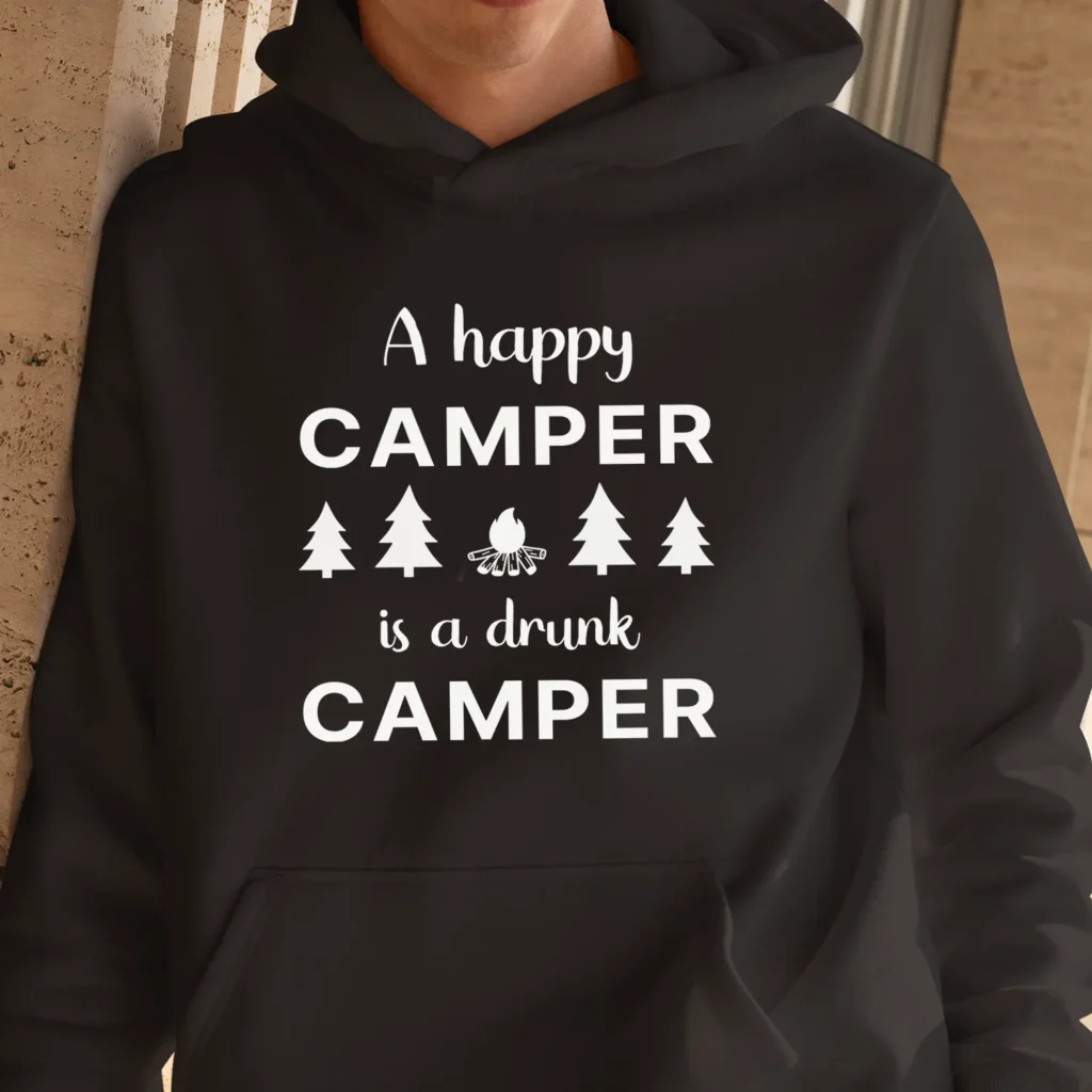 A man outside wearing a black Funny sweatshirt with an illustration of fur trees and a campfire saying "A happy camper is a drunk camper".