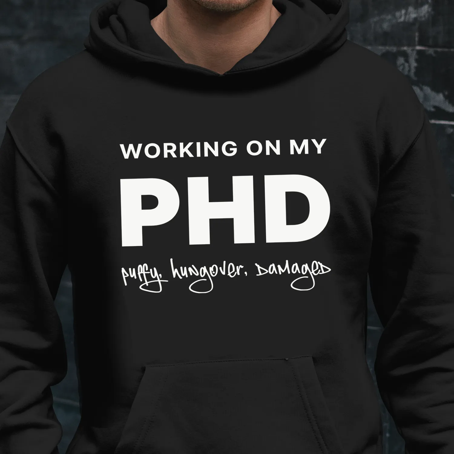 A man outside wearing a funny black hoodie saying "Working on my PHD: puffy, hungover, damaged".