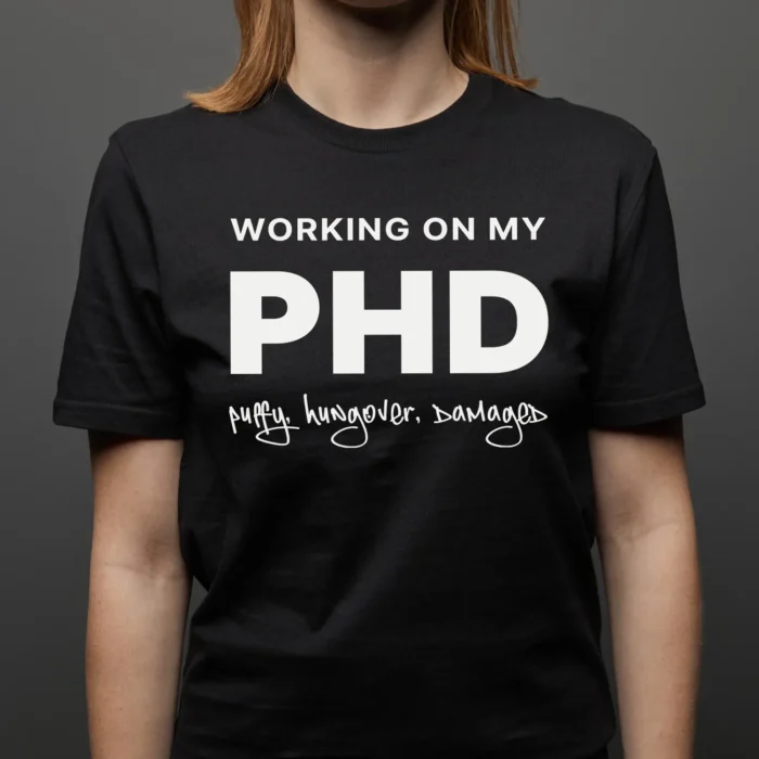 A young woman wearing a funny T-Shirt saying "Working on my PHD: puffy, hungover, damaged", Black Color