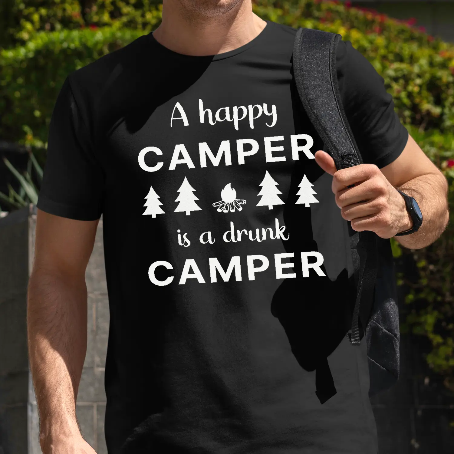 A man outside wearing a black Funny T-Shirt with an illustration of fur trees and a campfire saying "A happy camper is a drunk camper".