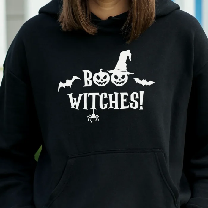 A young woman outside wearing a funny Halloween Hoodie with a witch's hat saying "Boo Witches!" Black color