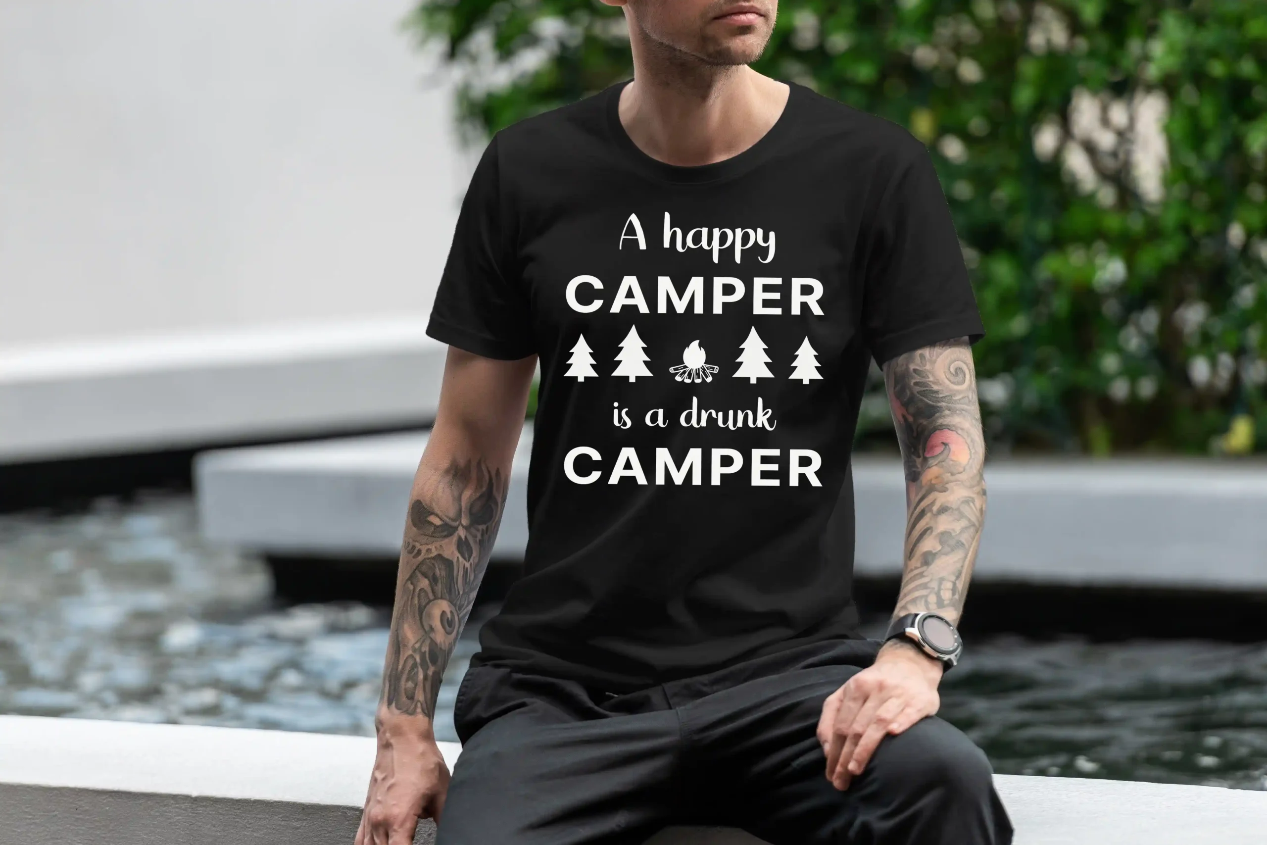 A man outside wearing a black Funny T-Shirt with an illustration of fur trees and a campfire saying "A happy camper is a drunk camper".