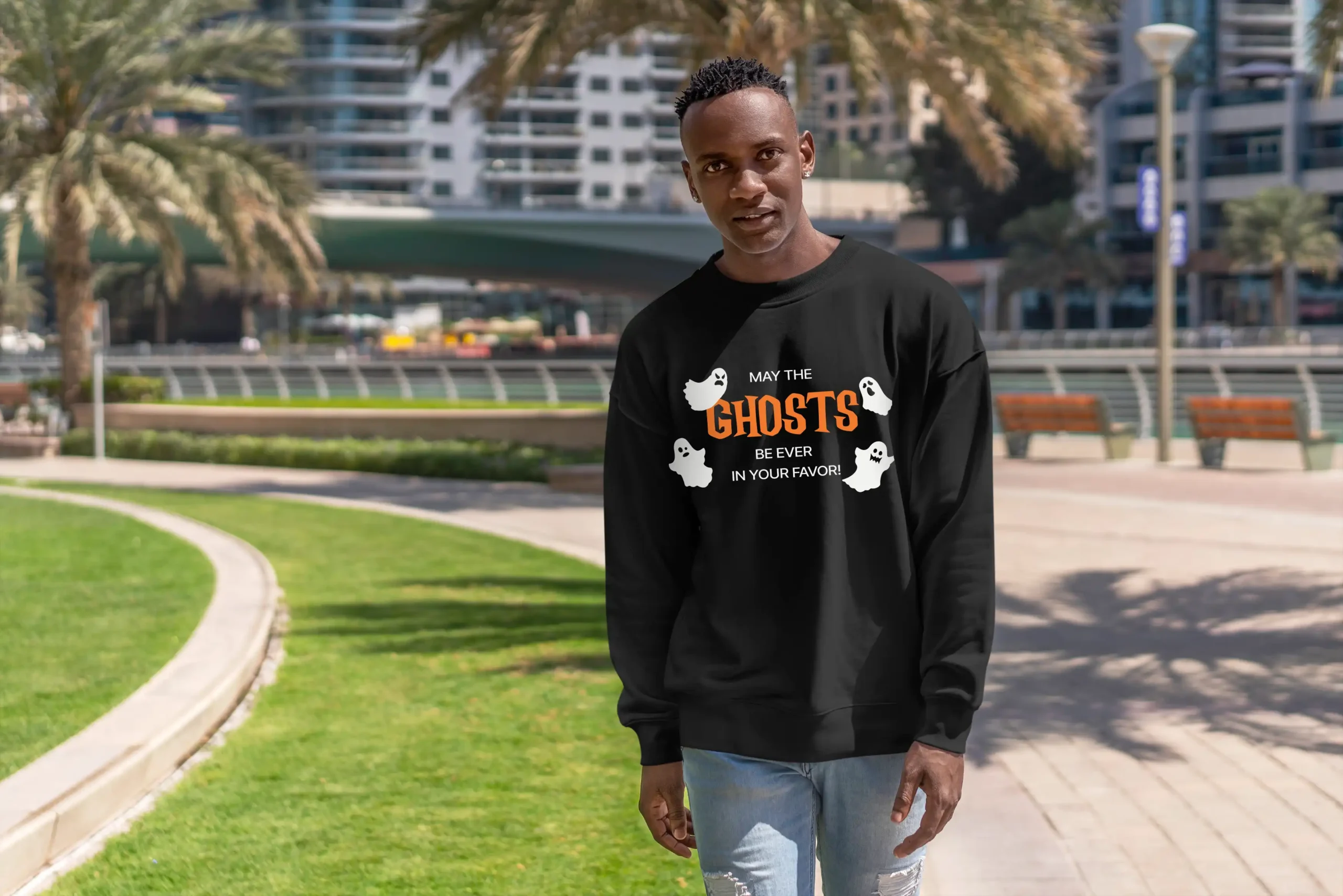A young african-american man outside wearing a funny Halloween Sweatshirt with ghosts saying "May the ghosts be ever in your favor!" Black color